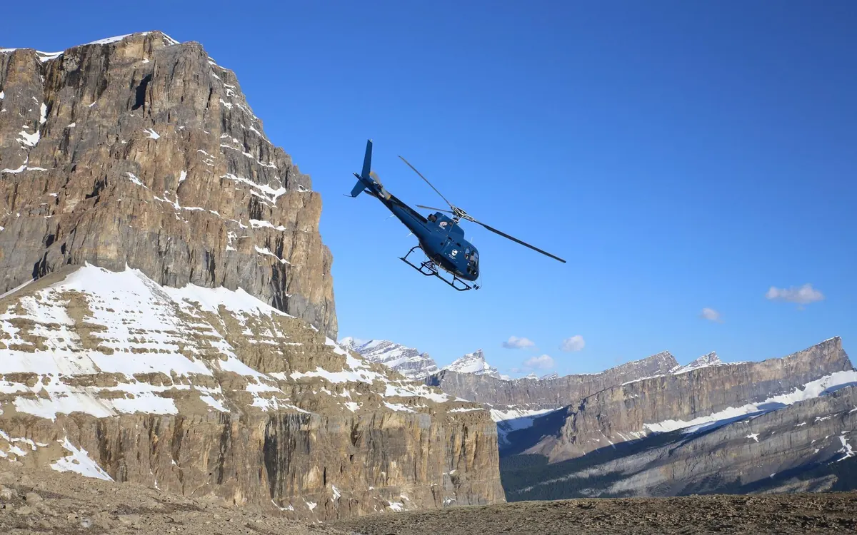 A helicopter beside a cliff above a turquoise lake in the Canadian Rockies